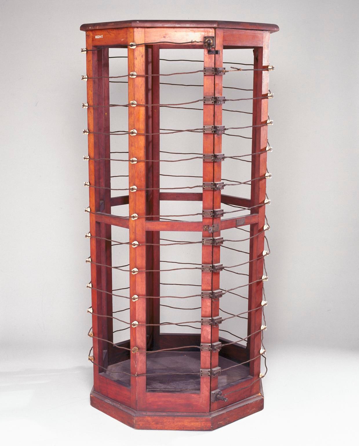 Figure 3 — The autoconduction solenoid itself, empty: a man-sized cage of turned wooden uprights wound with heavy wire, with a seat inside. There are no electrodes anywhere on it, and that is the whole idea —…