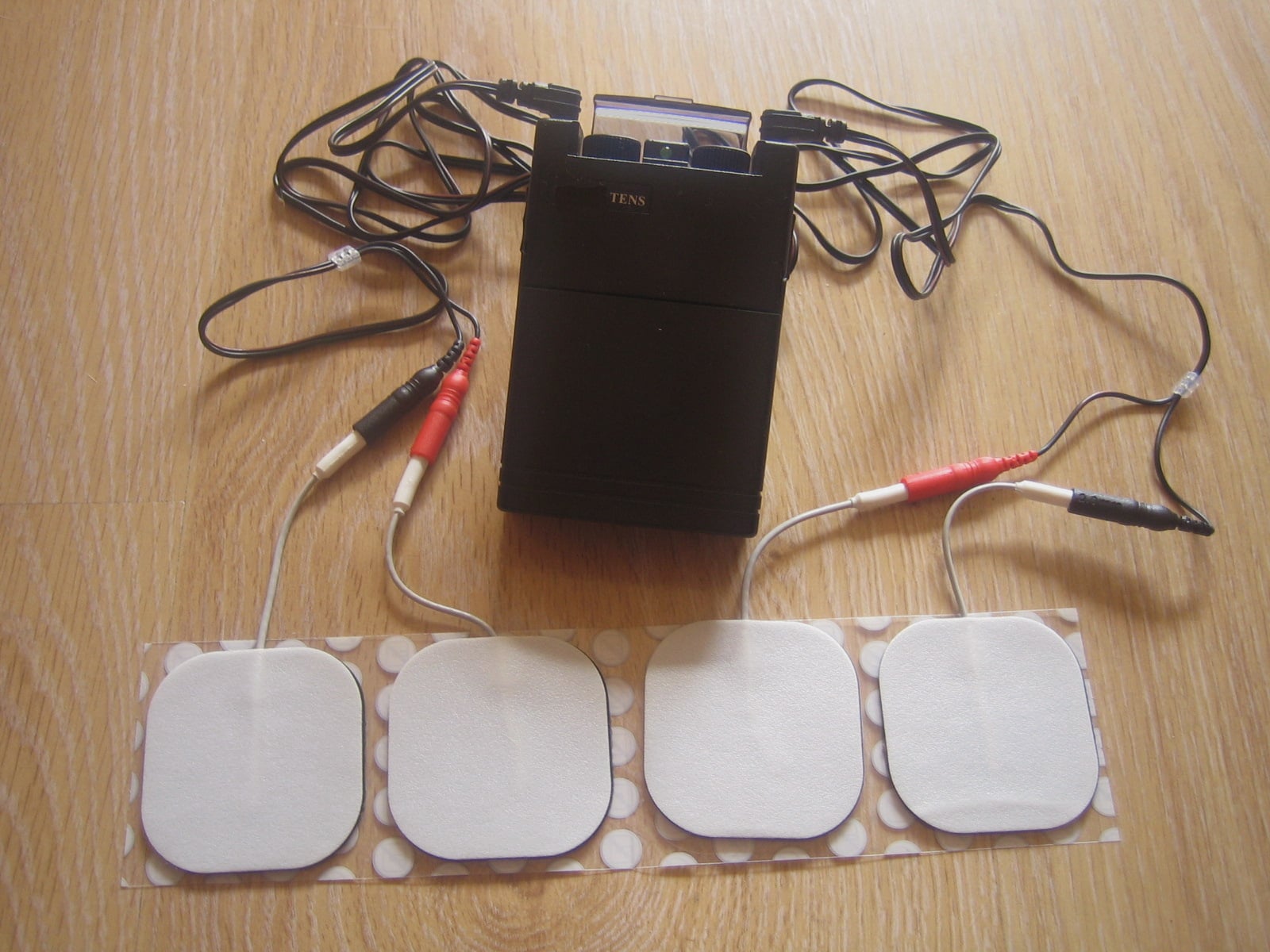 Figure 1 — A modern TENS unit: a pocket box of controls, lead wires, and four adhesive skin electrodes. Set it against a cased Victorian faradic battery and the family resemblance is uncomfortable — a current…