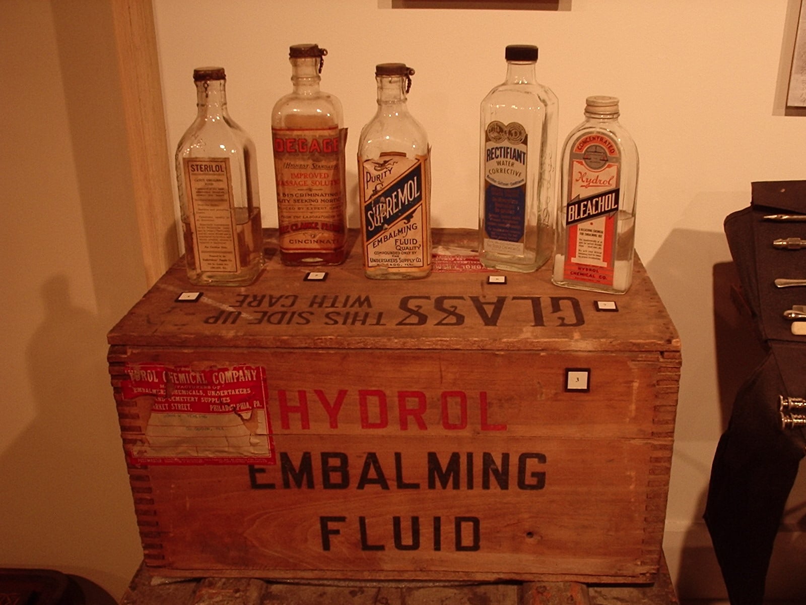 Figure 1 — A row of period fluid bottles standing on their own shipping crate — "HYDROL EMBALMING FLUID / GLASS — THIS SIDE UP — WITH CARE." Embossed and paper-labelled glass is how this chemistry survives to…