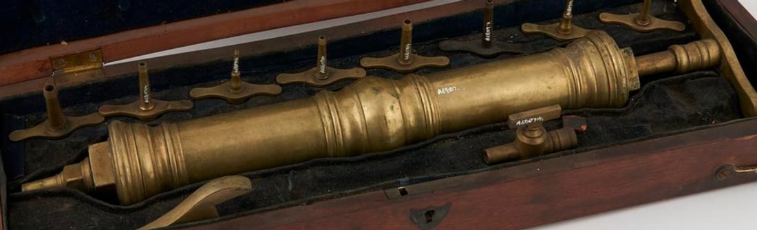 Figure 2 — A nickel-plated brass piston hand pump with its glass reservoir jar — the plunger and barrel visible above, the valved ports below; an object frequently likened to "a large syringe on a bottle."