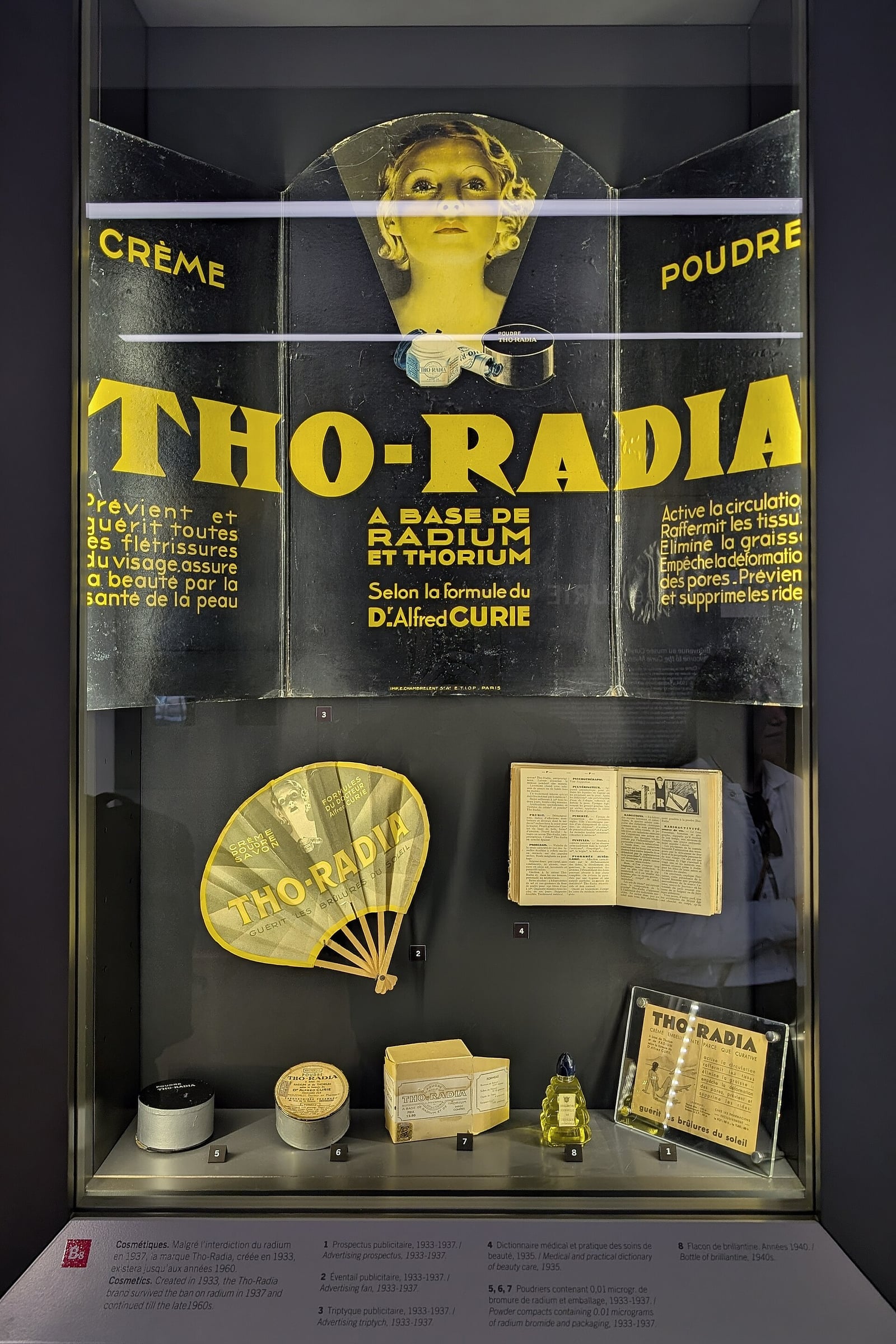 Figure 10 — The Tho-Radia line assembled — crème, poudre, the fan, the leaflets — in the Musée Curie. Radioactive cosmetics were not a fringe curiosity but a shelf-stocked consumer range with a house style.