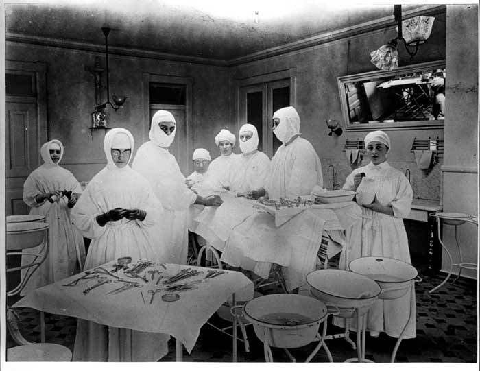 Figure 4 — The finished costume, about 1905: the whole team gowned, hooded, masked and gloved, working from draped instrument tables and enamelled basin stands. Nothing here is decorative — every element exis…